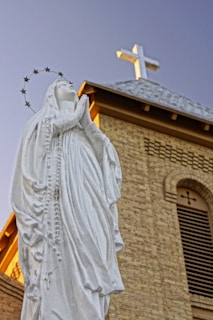 A white statue of a woman in prayer with a halo of stars around her head stands in front of a brick church building with a cross on the roof. The clear blue sky serves as the background.