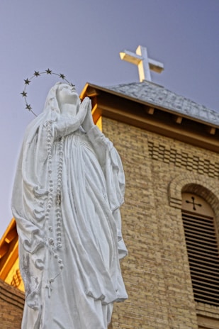 A white statue of a woman in prayer with a halo of stars around her head stands in front of a brick church building with a cross on the roof. The clear blue sky serves as the background.