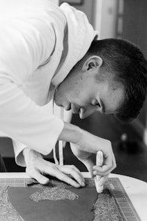 A person is focused on using a cutting tool on a piece of material laid out on a cutting mat. The person appears to be engaged in a crafting or design activity, wearing a light-colored hoodie. The image is in black and white, giving it a classic and timeless feel.