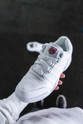 A person is holding a white athletic sneaker with a perforated design and a logo on the tongue. The shoe has white laces and silver eyelets, and it is set against a dark blurred background, highlighting the sneaker.