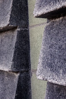 Close-up view of a textured wall composed of dark gray rectangular concrete blocks partially overlapping each other. The blocks are stacked in a staggered pattern creating a three-dimensional effect.