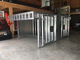 A metal storage or transport crate with a diamond plate pattern is situated in a garage. The crate is constructed from aluminum, featuring a sturdy frame with a grid-like design and handles on the sides.