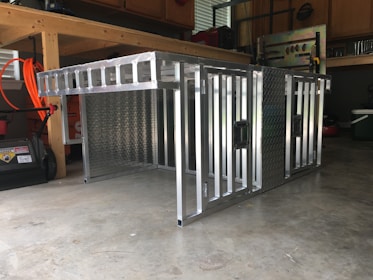 A metal storage or transport crate with a diamond plate pattern is situated in a garage. The crate is constructed from aluminum, featuring a sturdy frame with a grid-like design and handles on the sides.