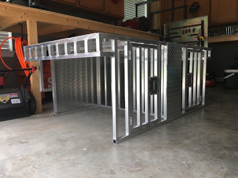 A metal storage or transport crate with a diamond plate pattern is situated in a garage. The crate is constructed from aluminum, featuring a sturdy frame with a grid-like design and handles on the sides.