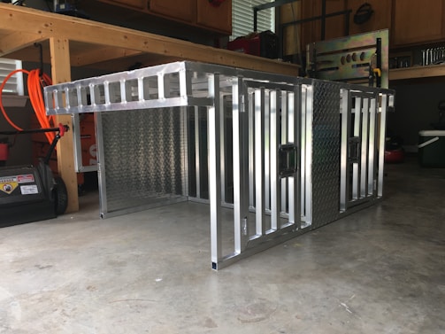 A metal storage or transport crate with a diamond plate pattern is situated in a garage. The crate is constructed from aluminum, featuring a sturdy frame with a grid-like design and handles on the sides.