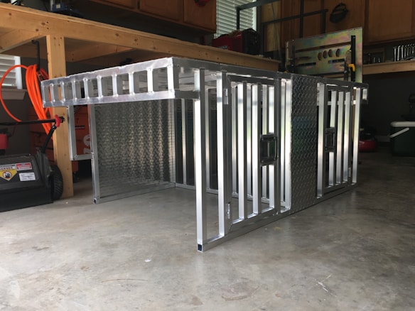 A metal storage or transport crate with a diamond plate pattern is situated in a garage. The crate is constructed from aluminum, featuring a sturdy frame with a grid-like design and handles on the sides.