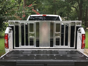 A white pickup truck with an open tailgate is parked outdoors; a metallic, cage-like structure is fitted onto the truck bed. The structure includes a diamond-plate panel and several barred compartments. Surrounding the truck, there are trees and bushes with green leaves and some red flowers.