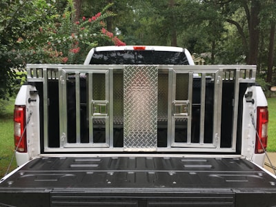 A white pickup truck with an open tailgate is parked outdoors; a metallic, cage-like structure is fitted onto the truck bed. The structure includes a diamond-plate panel and several barred compartments. Surrounding the truck, there are trees and bushes with green leaves and some red flowers.
