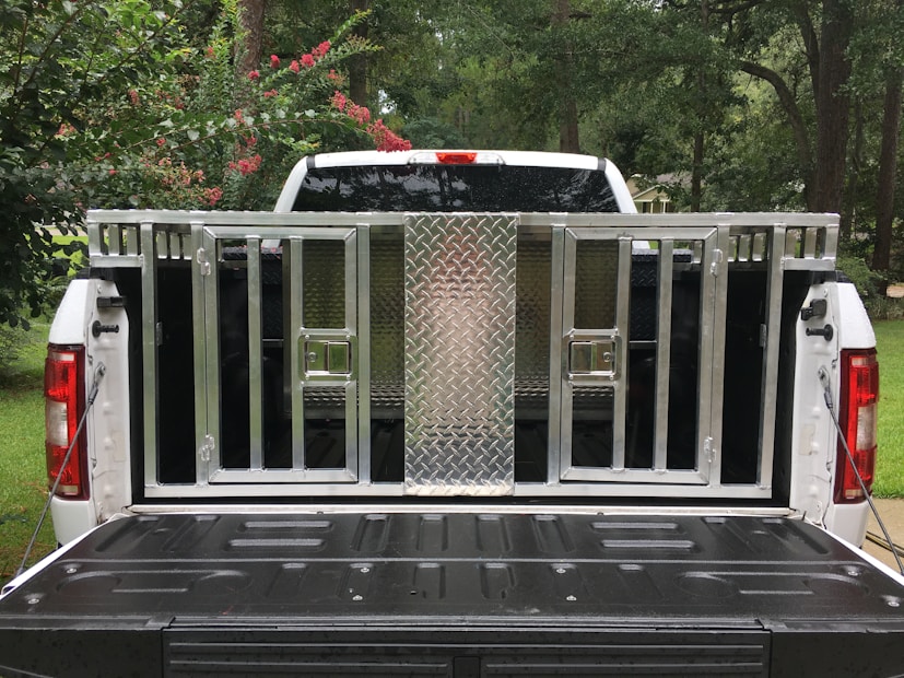 A white pickup truck with an open tailgate is parked outdoors; a metallic, cage-like structure is fitted onto the truck bed. The structure includes a diamond-plate panel and several barred compartments. Surrounding the truck, there are trees and bushes with green leaves and some red flowers.