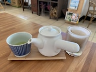 A white teapot is placed on a wooden tray alongside a small blue-striped cup containing green tea and a double-walled glass cup with tea inside. The setting is a cozy room with a wooden table and a tiled floor, with some furniture and decorative items visible in the background.