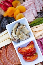 A charcuterie board featuring an assortment of foods, including sliced strawberries, dried apricots, figs, and salami. There is also a section with cheese slices and cured meat. Centrally placed is a dish with marinated artichoke hearts and roasted red peppers.