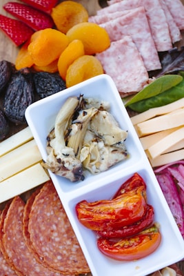 A charcuterie board featuring an assortment of foods, including sliced strawberries, dried apricots, figs, and salami. There is also a section with cheese slices and cured meat. Centrally placed is a dish with marinated artichoke hearts and roasted red peppers.