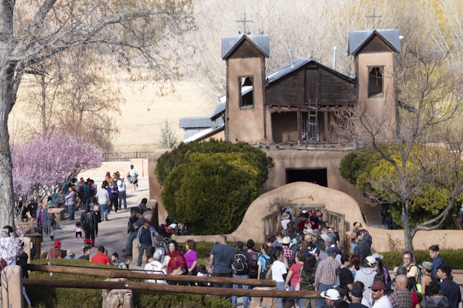 A large group of people is gathered outside a rustic adobe-style church with two towers. The scene is lively, with people walking towards the entrance, surrounded by bare and flowering trees. The setting appears to be in a rural area during a clear, sunny day.