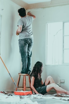 Two people are engaged in painting a room. One person stands on a stool and uses a brush to paint the upper part of the wall, while the other person sits on the floor beside a paint roller and tray. The room appears well-lit through a nearby window.