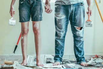 Two individuals stand barefoot on a floor covered with newspapers, holding paintbrushes in their hands. Their clothes are splattered with paint, indicating they have been painting. One person is wearing denim shorts, and the other is wearing ripped jeans. A paint roller with a red handle is on the floor, suggesting an ongoing painting project.