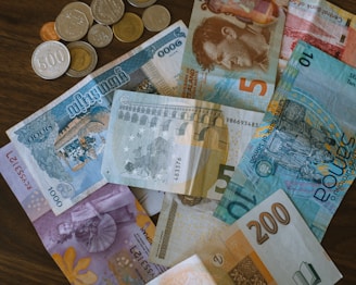 A collection of various currency bills and coins are scattered on a wooden surface, showcasing different denominations and designs from multiple countries.