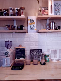 A countertop setup in a cozy kitchen with various coffee-making items and decor. Shelves hold jars with coffee beans and kitchen utensils. A grinder, scale, and several glasses of coffee are placed on the counter. Decorative signs with motivational quotes and coffee themes are displayed.
