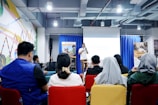 A speaker stands at the front of a room filled with people seated in colorful chairs, giving a presentation. The setting appears to be a modern seminar or workshop with banners displayed on either side of a projection screen. The walls have vibrant geometric designs, and the environment is well-lit with overhead lights.