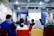 A speaker stands at the front of a room filled with people seated in colorful chairs, giving a presentation. The setting appears to be a modern seminar or workshop with banners displayed on either side of a projection screen. The walls have vibrant geometric designs, and the environment is well-lit with overhead lights.