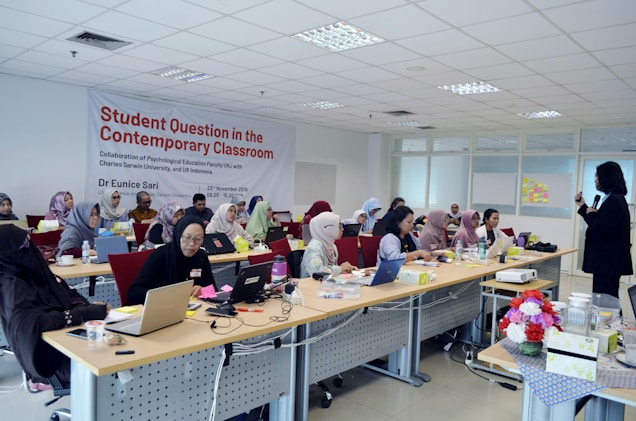 A classroom setting with several people seated at long tables with laptops and papers, attending a presentation or workshop. A presenter is standing to the right of the image, engaging with the audience. The room is well-lit, with a large banner on the wall displaying the topic 'Student Question in the Contemporary Classroom'. Various items such as flowers, cups, and boxes are placed on the tables.