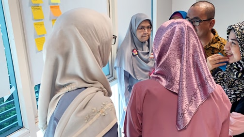 A group of people are gathered around a whiteboard with colorful sticky notes. The individuals are dressed in various patterns and colors, including hijabs. They appear to be engaged in a discussion or brainstorming session.