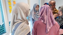 A group of people are gathered around a whiteboard with colorful sticky notes. The individuals are dressed in various patterns and colors, including hijabs. They appear to be engaged in a discussion or brainstorming session.