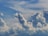 Massive white cumulus clouds are dispersed across a deep blue sky, creating a dynamic and textured appearance. The clouds have varied shapes and are illuminated by sunlight, giving them soft gradients of light and shadow.