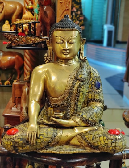A golden Buddha statue intricately detailed with ornate patterns, seated in a meditative posture. The statue has colored embellishments, including red and blue stones. Surrounding the statue are wooden sculptures and decorative artifacts, creating an atmosphere of cultural significance.
