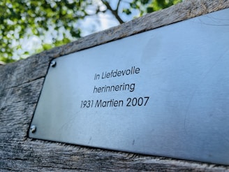 A metal plaque is mounted on a weathered wooden surface, bearing the inscription 'In Liefdevolle herinnering 1931 Martien 2007' in a serif font. The background consists of blurred green foliage, suggesting an outdoor setting.