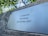 A metal plaque is mounted on a weathered wooden surface, bearing the inscription 'In Liefdevolle herinnering 1931 Martien 2007' in a serif font. The background consists of blurred green foliage, suggesting an outdoor setting.