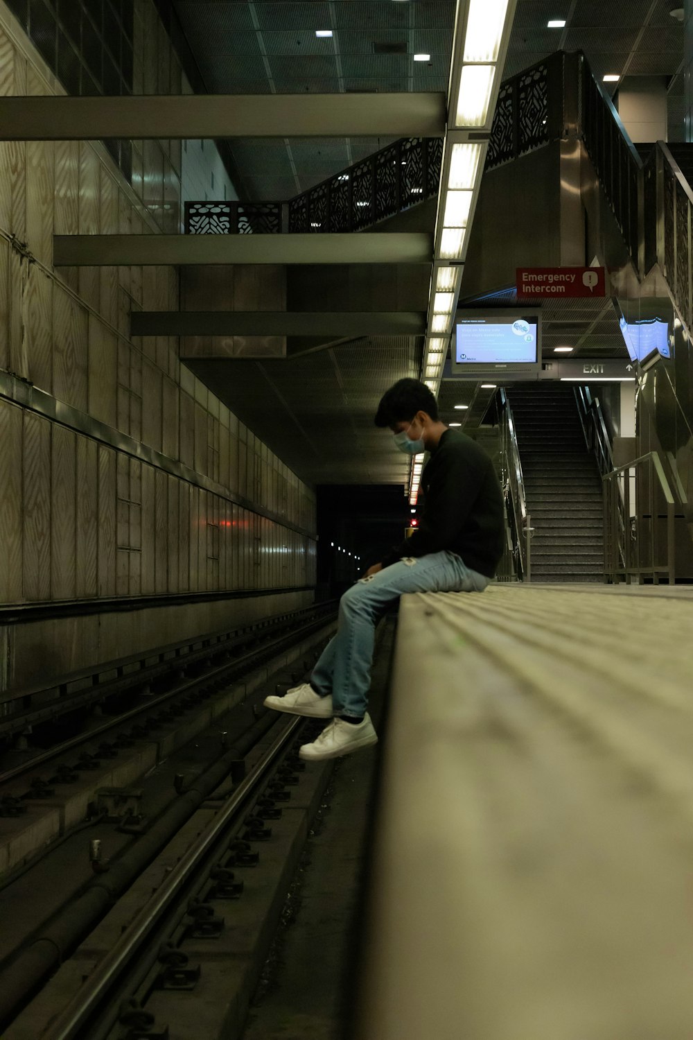 man in black jacket and blue denim jeans sitting on train rail
