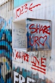 Graffiti-covered wall featuring bold red text with phrases such as '1023', 'SHARE', and 'YOU ARE LOVE'. The markings have a street art style, with a mix of blue and black paint in the background.