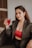 A woman sitting in a wicker chair, wearing a dark suit and a red lace top, is holding a glass with a red drink. Her long brown hair is loose, and she has sunglasses resting on her head. The setting appears to be indoors with a neutral-toned wall in the background.