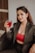 A woman sitting in a wicker chair, wearing a dark suit and a red lace top, is holding a glass with a red drink. Her long brown hair is loose, and she has sunglasses resting on her head. The setting appears to be indoors with a neutral-toned wall in the background.
