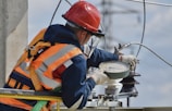 A worker wearing a red hard hat and orange safety vest is adjusting equipment on an electrical pole. The individual is equipped with protective gloves and harness, and is focused on a round device attached to the structure. Power lines and a blue sky with clouds form the background.