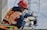 A worker wearing a red hard hat and orange safety vest is adjusting equipment on an electrical pole. The individual is equipped with protective gloves and harness, and is focused on a round device attached to the structure. Power lines and a blue sky with clouds form the background.