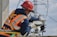 A worker wearing a red hard hat and orange safety vest is adjusting equipment on an electrical pole. The individual is equipped with protective gloves and harness, and is focused on a round device attached to the structure. Power lines and a blue sky with clouds form the background.