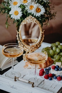 Two elegant champagne glasses filled with a light-colored beverage are placed on a decorative white tray. A small bouquet of white daisies with yellow centers sits beside a gold ornate mirror. On the tray, there's a croissant, grapes, raspberries, blueberries, and cheese, creating a luxurious and inviting brunch setting.