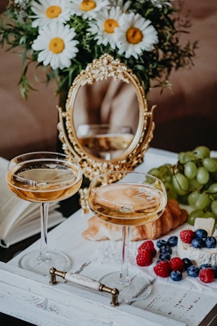 Two elegant champagne glasses filled with a light-colored beverage are placed on a decorative white tray. A small bouquet of white daisies with yellow centers sits beside a gold ornate mirror. On the tray, there's a croissant, grapes, raspberries, blueberries, and cheese, creating a luxurious and inviting brunch setting.