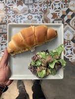 A hand is holding a square plate containing a croissant sandwich filled with vegetables and a side salad. The salad consists of mixed greens and croutons. The background features a colorful, patterned tile floor.