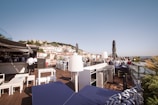 An outdoor rooftop terrace with a panoramic view of a cityscape under a clear blue sky. Several people are seated at tables, enjoying the view or socializing. The area features modern outdoor furniture, including tables and chairs, with some shade provided by large umbrellas. A bar area is visible with staff members present, and lush greenery adds a touch of nature.