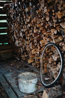 A neatly stacked pile of cut firewood fills the right half of the scene, with a variety of logs organized tightly together. A worn metal bucket sits on the ground in front of the logs, showing signs of rust and use. Next to the bucket, an old bicycle tire leans against the wood pile. The setting appears rustic, with a wooden platform and scattered wood debris creating a rough texture on the floor.