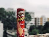 A red cylindrical can of Pringles with the iconic logo featuring a stylized man's face and the word 'Original' prominently displayed. The background shows an out-of-focus urban area with buildings and trees.