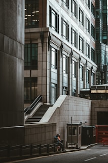 The photograph captures a modern urban landscape featuring a large, multi-story building with numerous windows and intricate architectural details. On the street level, a pedestrian dressed in casual clothing leans against a low wall near a set of stairs. The scene has a muted color palette, typical of an overcast day in a city environment.