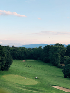 A scenic landscape featuring a golf course surrounded by dense trees, with a manicured green and sand bunkers. The horizon shows distant mountains under a clear blue sky with a few pink clouds. Birds are flying in the sky, adding a tranquil element to the scene.