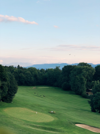 A scenic landscape featuring a golf course surrounded by dense trees, with a manicured green and sand bunkers. The horizon shows distant mountains under a clear blue sky with a few pink clouds. Birds are flying in the sky, adding a tranquil element to the scene.