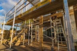 A detailed construction scene involving a complex arrangement of metal scaffolding and ladders under a clear sky. The scaffolding is marked with branding and there are several vertical and horizontal support structures. A wooden beam and some construction materials are visible, with sunlight creating shadows across the scene.