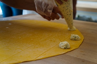A close-up view of hands using a piping bag to place a creamy, herb-speckled filling onto a sheet of yellow pasta dough. The dough is scored into a grid, indicating preparation for making stuffed pasta such as ravioli. The setting appears to be a kitchen with wooden surfaces, suggesting a homemade or artisanal cooking process.