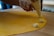 A close-up view of hands using a piping bag to place a creamy, herb-speckled filling onto a sheet of yellow pasta dough. The dough is scored into a grid, indicating preparation for making stuffed pasta such as ravioli. The setting appears to be a kitchen with wooden surfaces, suggesting a homemade or artisanal cooking process.