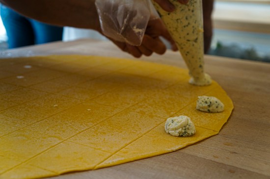 A close-up view of hands using a piping bag to place a creamy, herb-speckled filling onto a sheet of yellow pasta dough. The dough is scored into a grid, indicating preparation for making stuffed pasta such as ravioli. The setting appears to be a kitchen with wooden surfaces, suggesting a homemade or artisanal cooking process.